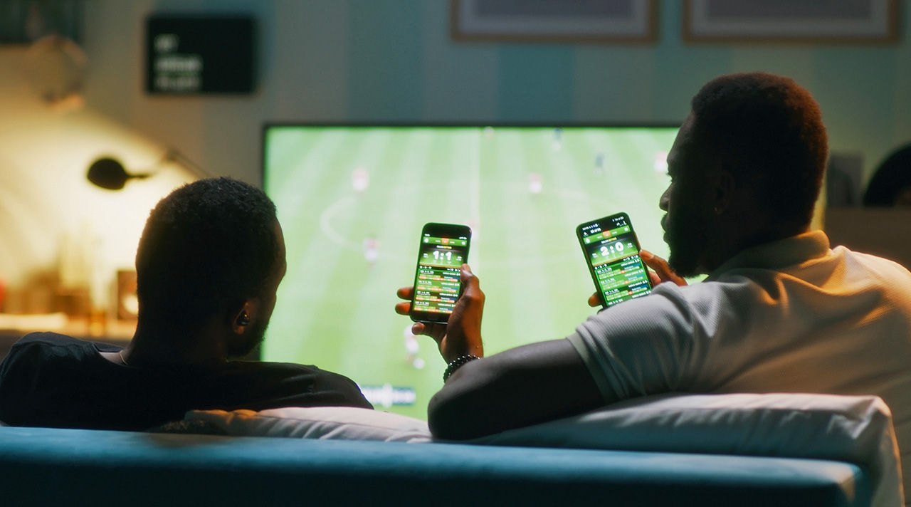 Two people on a couch watching a sporting event while also on their smart phones using the sports streaming app. 